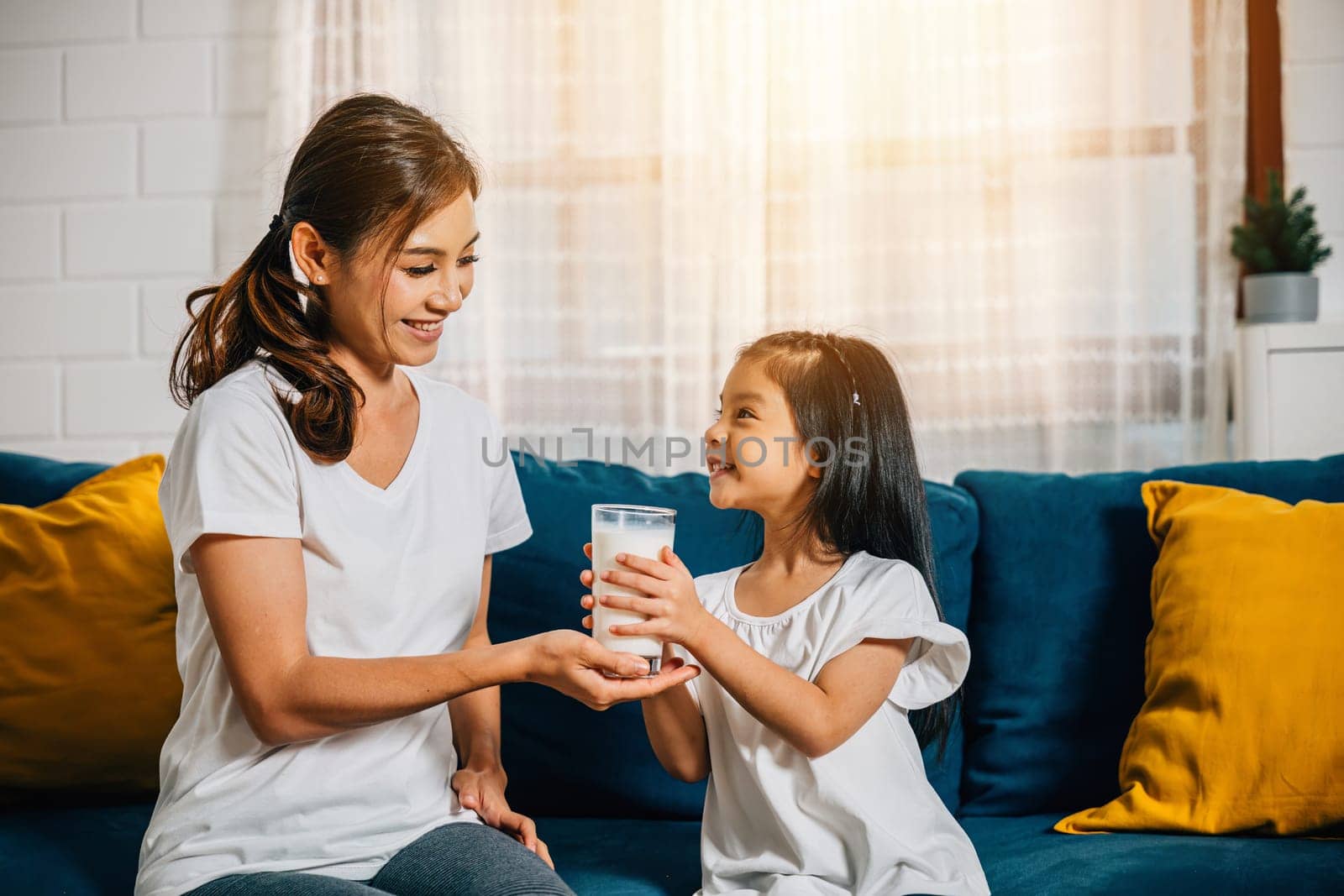 A joyful scene as an Asian mother gives her daughter a glass of milk on the living room sofa. This image radiates affection innocence and the happiness of family breakfasts.