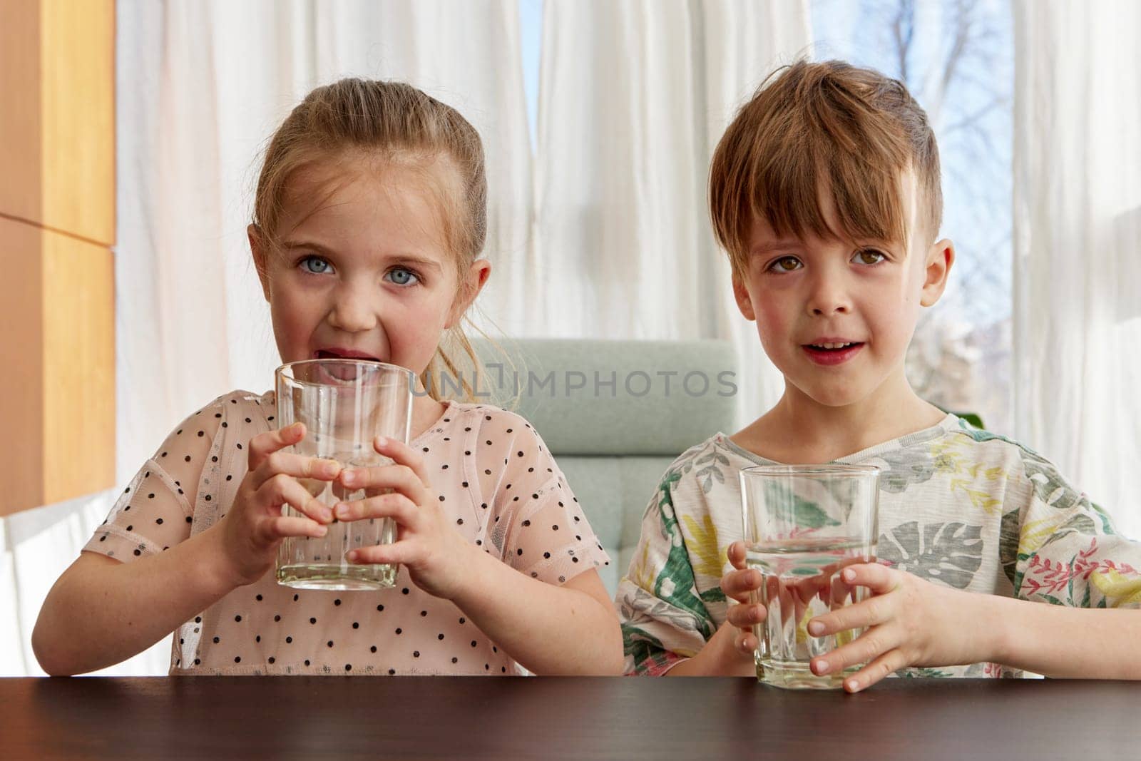 Brother and sister sit at kitchen table drinking water from glasses. Children quench thirst before going to school in morning closeup slow motion