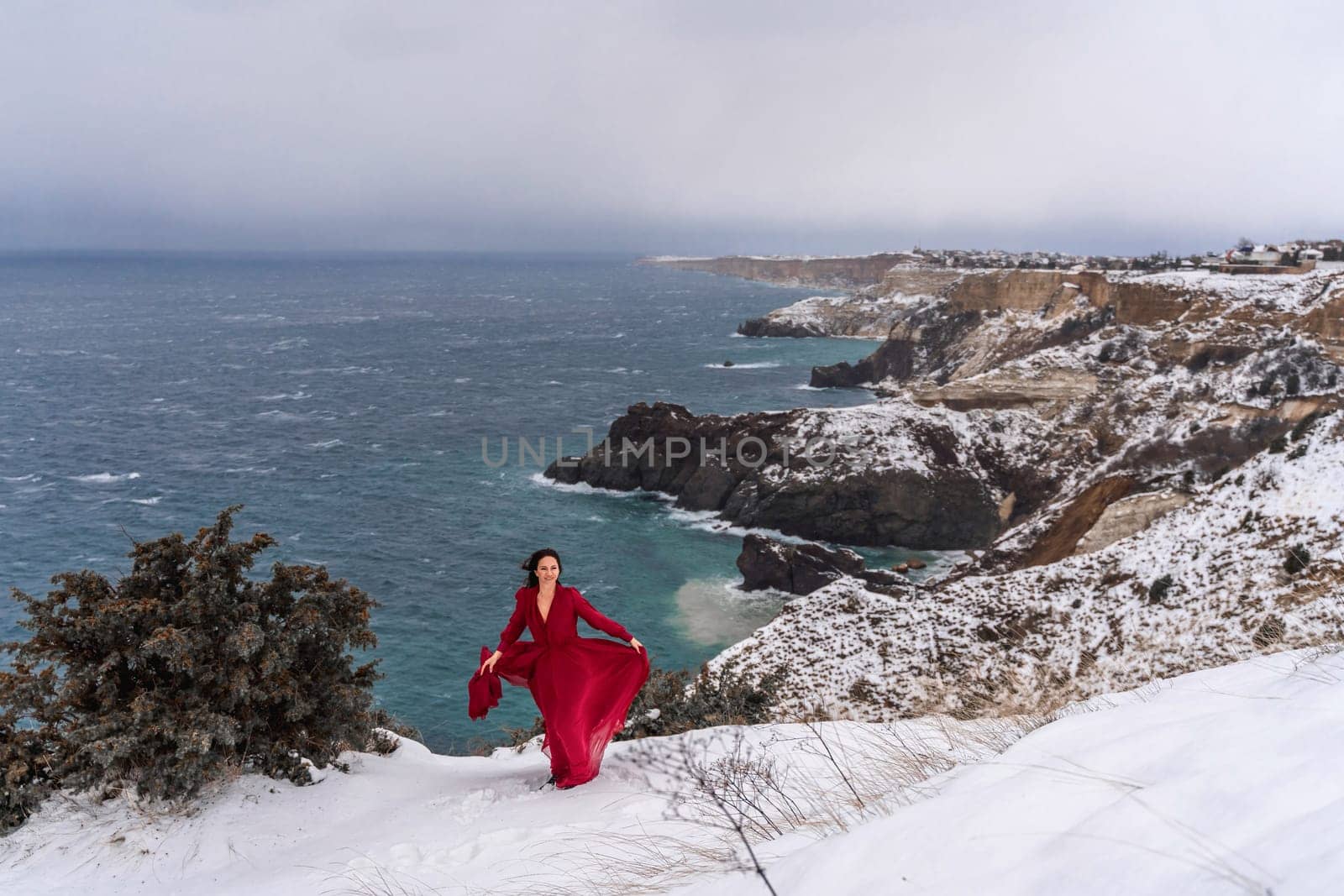 Woman red dress snow sea. Happy woman in a red dress in the snowy mountains by the emerald sea. The wind blows her clothes, posing against sea and snow background. by Matiunina