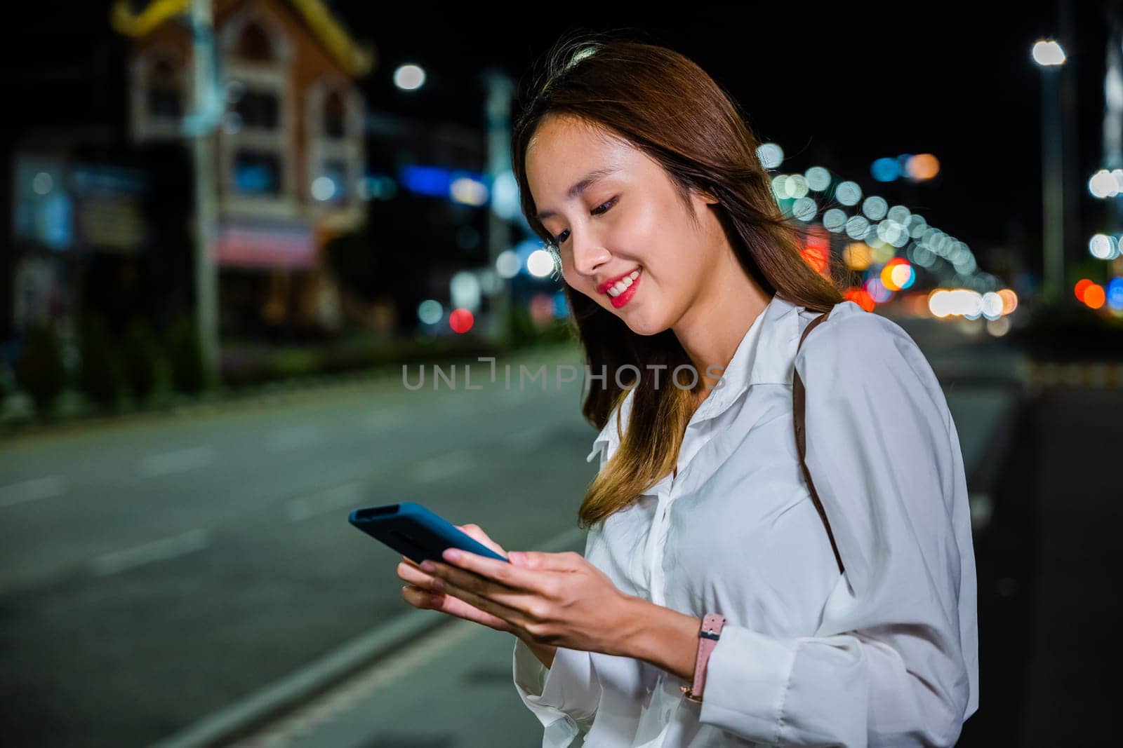 A woman stands on the sidewalk at night, holding her cellphone while waiting for transportation, with a vibrant orange and teal color scheme.