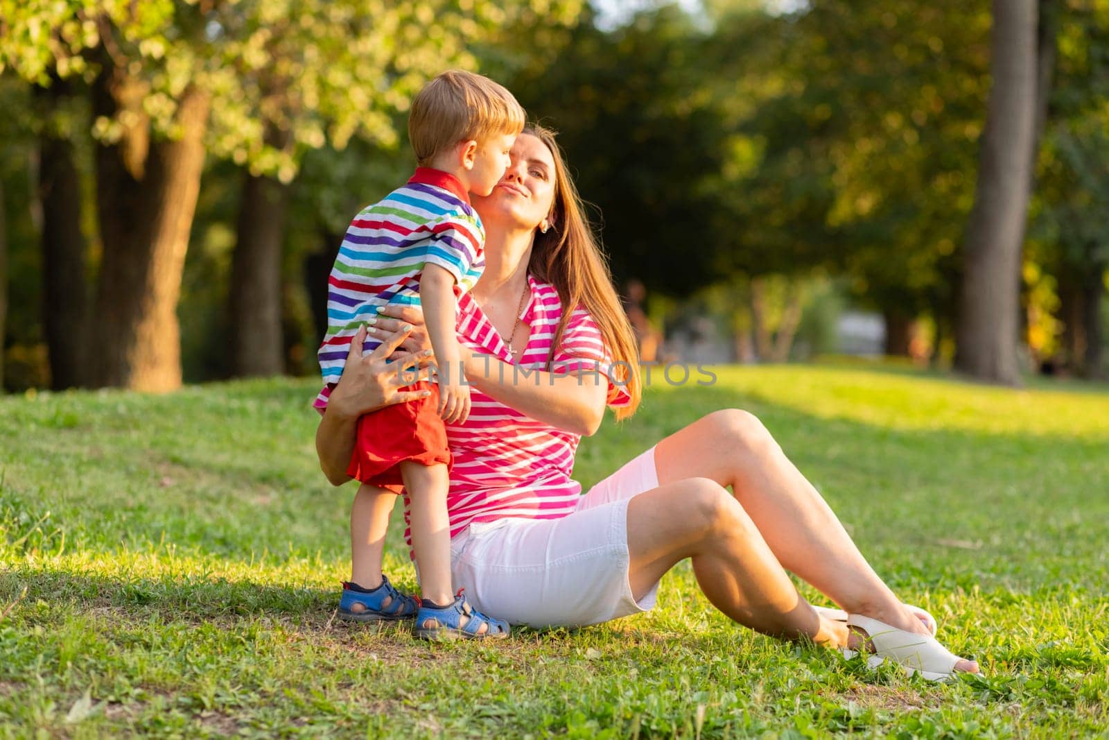 A tender moment as a mother embraces her young son in a loving hug while sitting in a sunlit park, enjoying the outdoors.