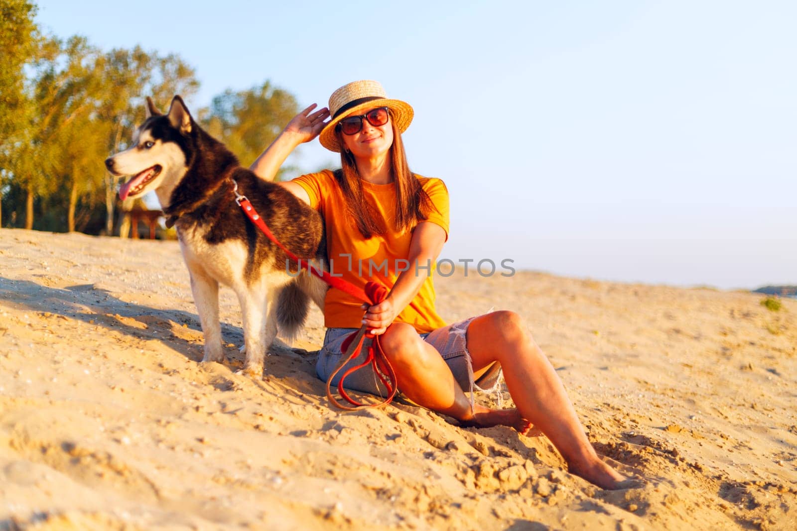A woman in a straw hat and sunglasses relaxes on the beach with her Siberian Husky, enjoying a beautiful sunset together.