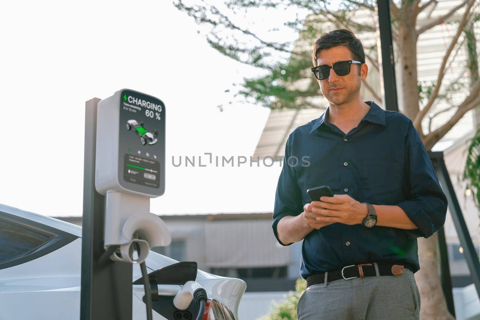 Young man use smartphone to pay for electricity at public EV car charging station at city commercial mall parking lot. Modern environmental and sustainable urban lifestyle with EV vehicle. Expedient