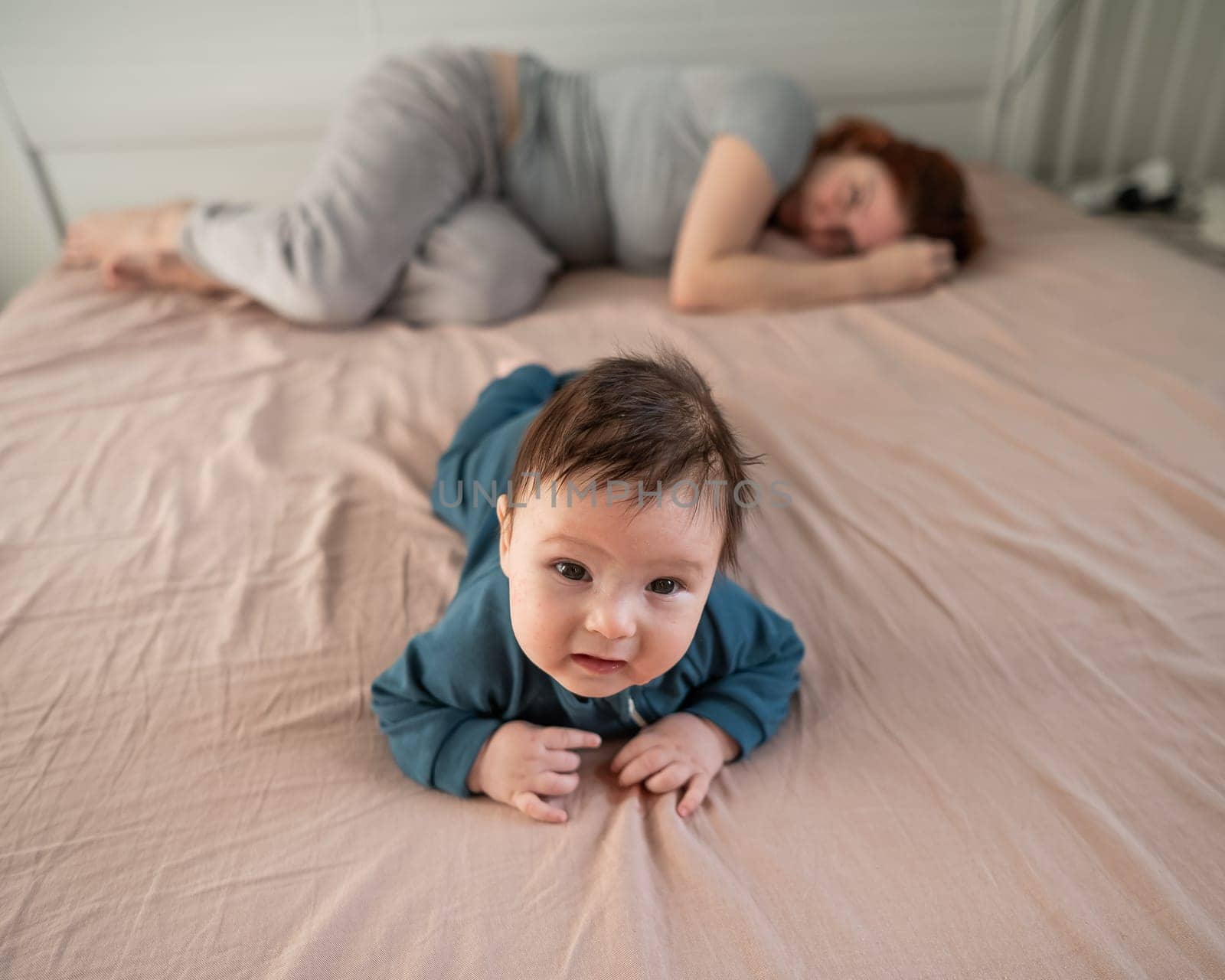 A three-month-old boy lies on his stomach on the bed and his mother sleeps behind him. Postpartum depression. by mrwed54