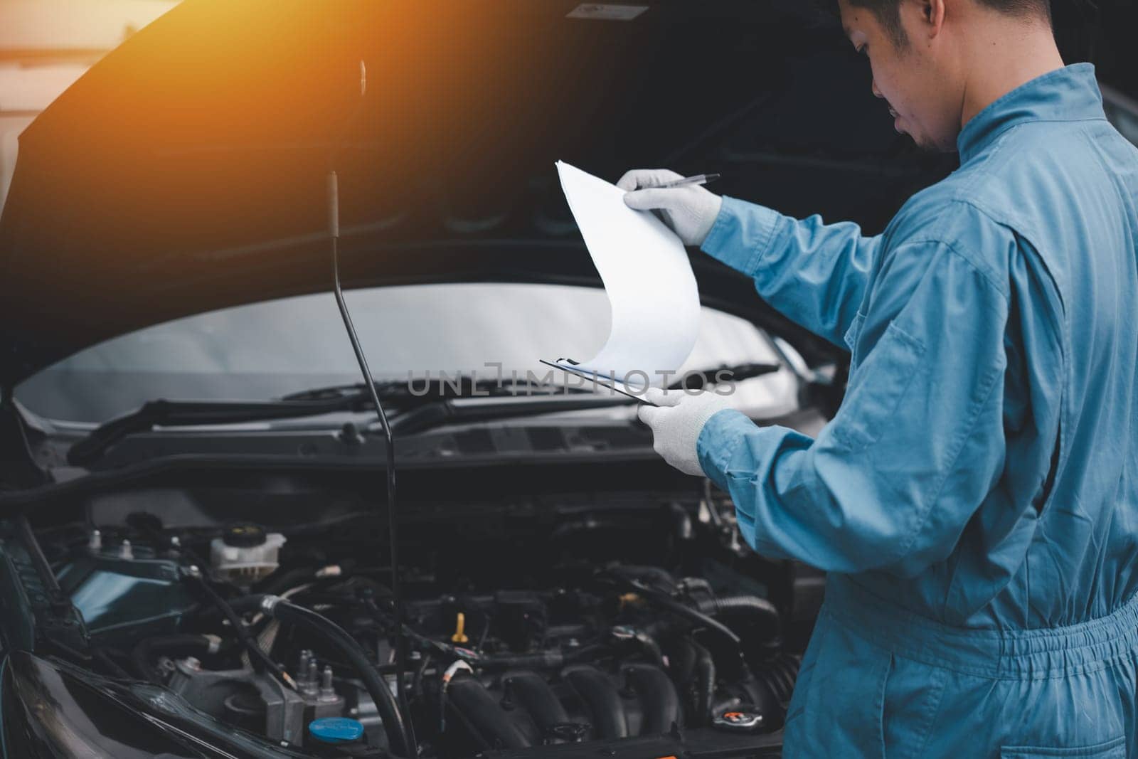 Asian mechanic in blue workwear writing on a paper, while repairing a car in an auto repair shop. Close-up shot of hand.