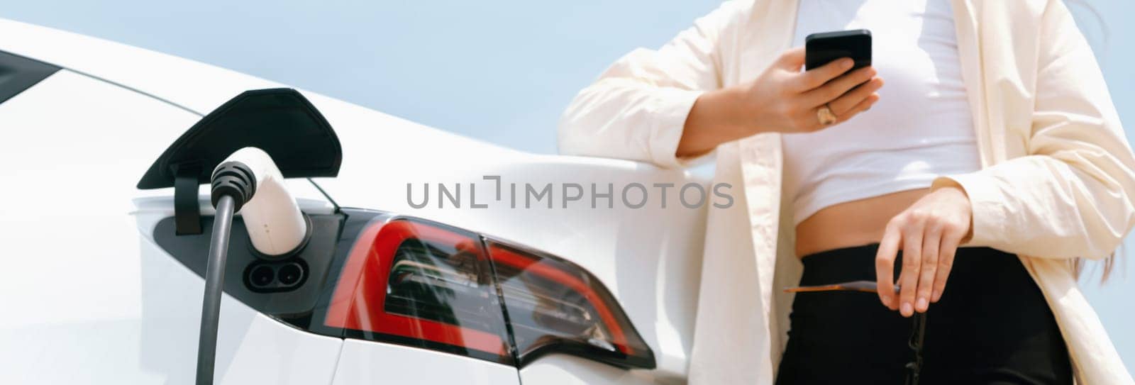 Young woman use smartphone to pay for electricity at public EV car charging station green city park. Modern environmental and sustainable urban lifestyle with EV vehicle. Panorama Expedient