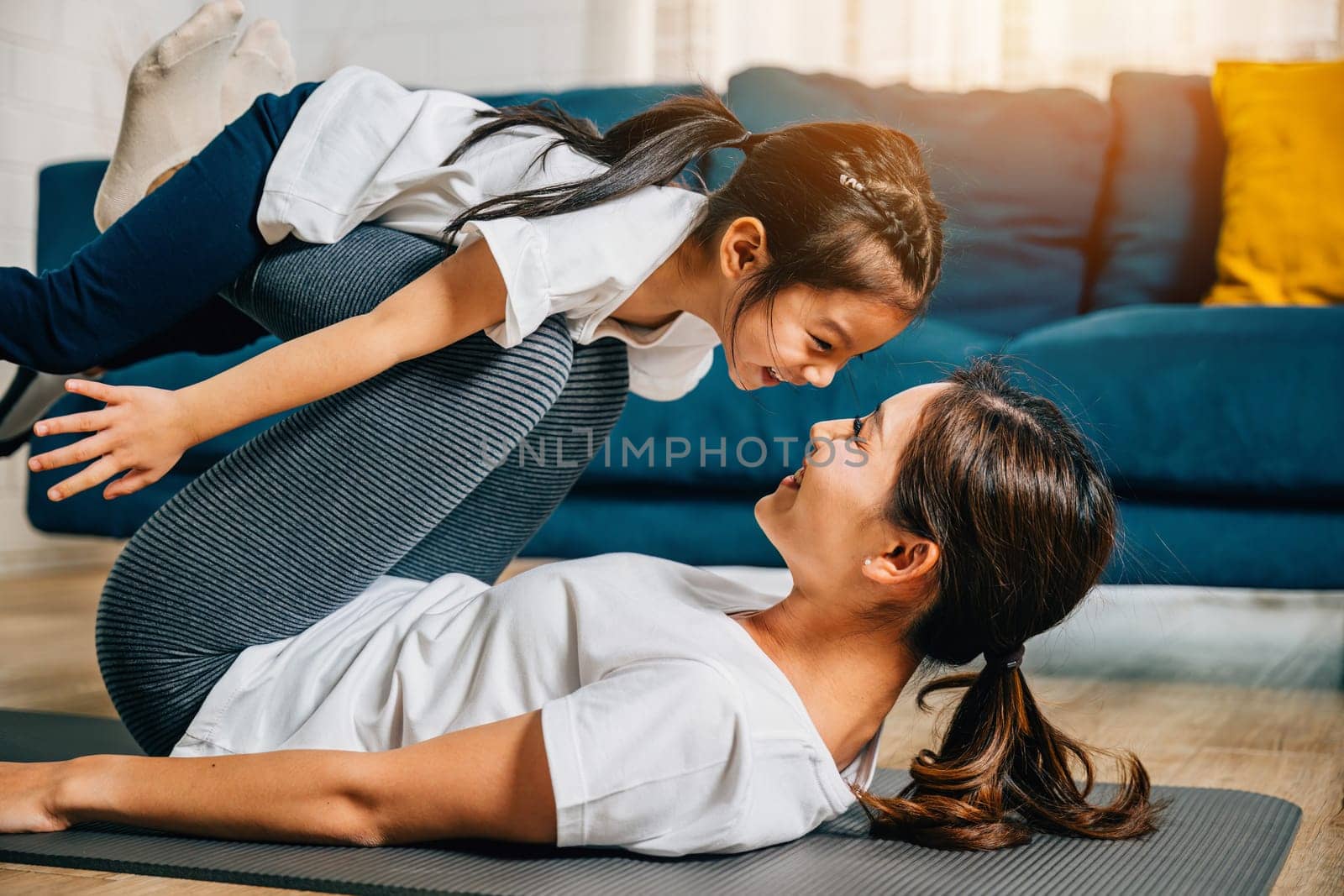 A mother holds her child in a playful family fitness pose imagining themselves 'flying' like airplanes during their Pilates and yoga routine promoting trust harmony and happiness.