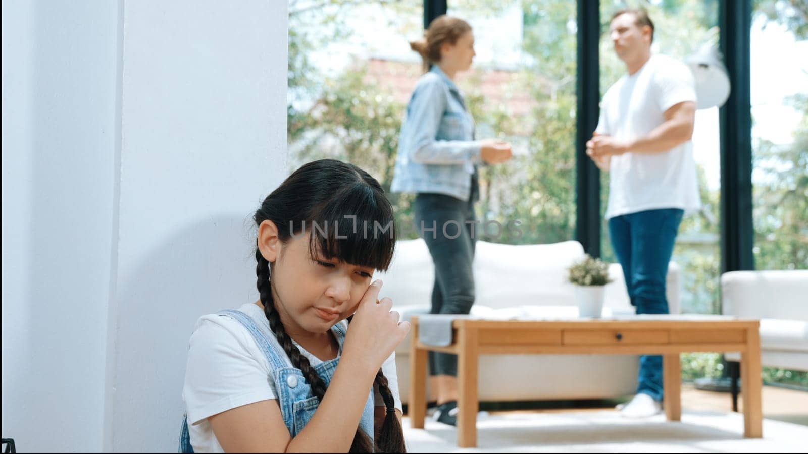 Stressed and unhappy young girl huddle in corner, cover her ears blocking sound of her parent arguing in background. Domestic violence at home and traumatic childhood develop to depression. Synchronos