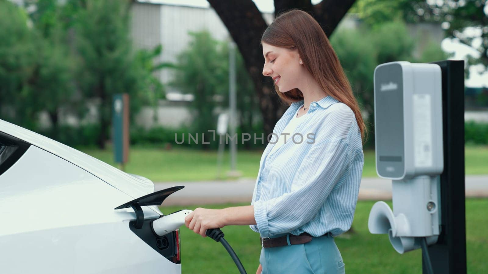 Young woman recharging battery for electric car during road trip travel EV car in natural forest or national park. Eco friendly travel during vacation and holiday. Exalt