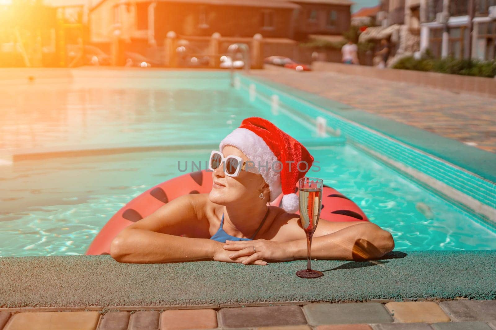 Woman pool Santa hat. A happy woman in a blue bikini, a red and white Santa hat and sunglasses poses near the pool with a glass of champagne standing nearby. Christmas holidays concept