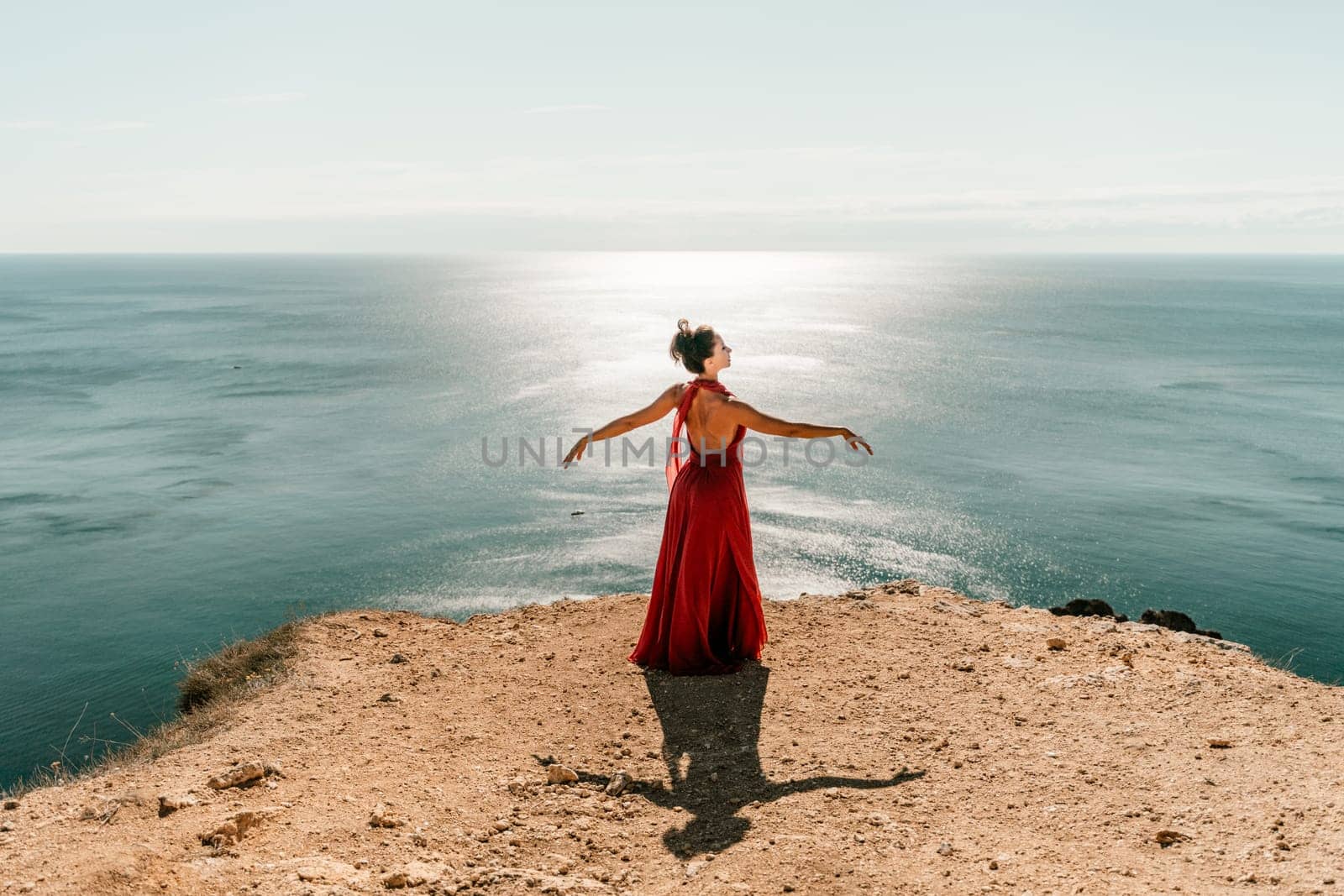 Woman red dress sea. posing on a rocky outcrop high above the sea. Girl on the nature on blue sky background. Fashion photo. by Matiunina