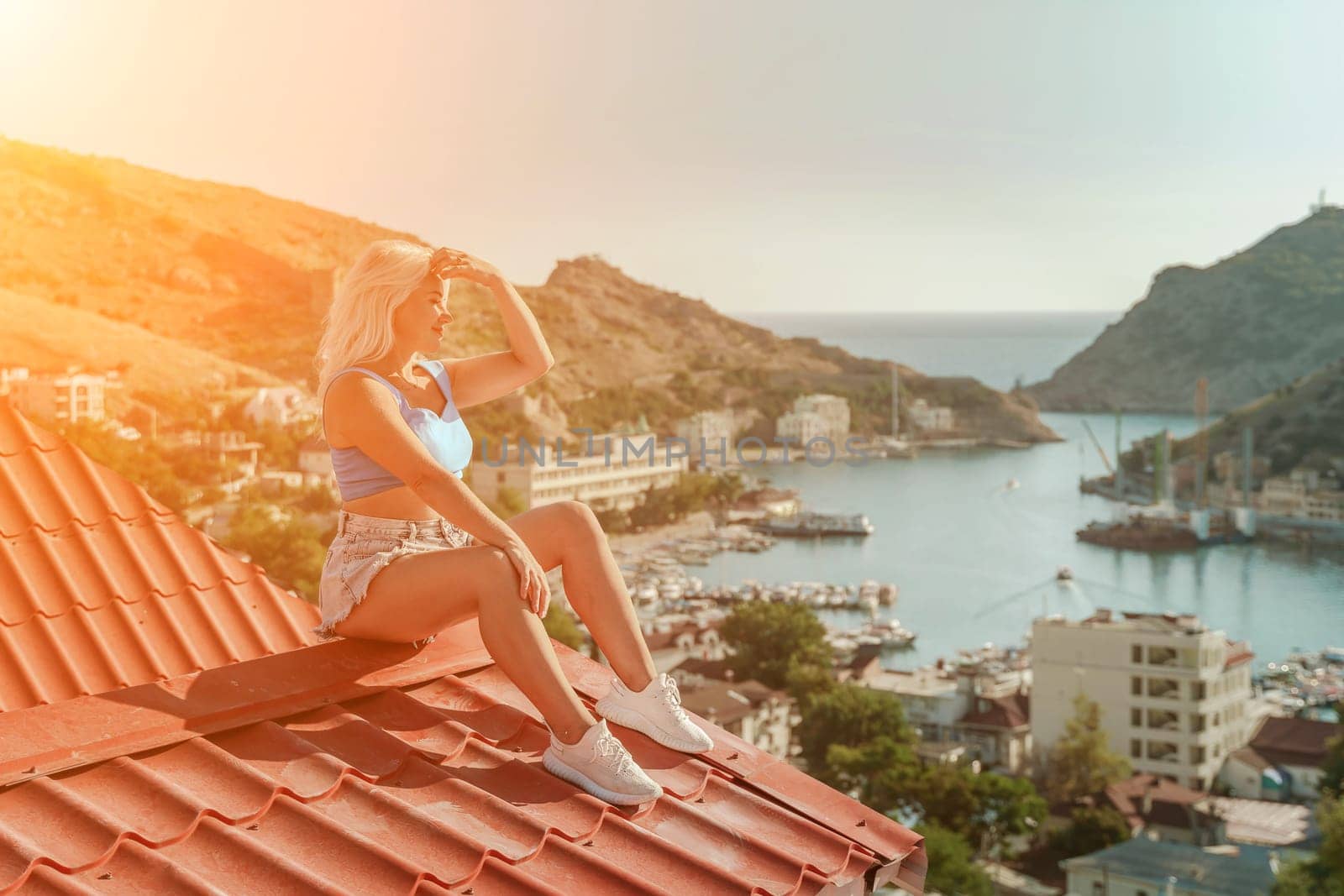 Woman sits on rooftop, enjoys town view and sea mountains. Peaceful rooftop relaxation. Below her, there is a town with several boats visible in the water. Rooftop vantage point