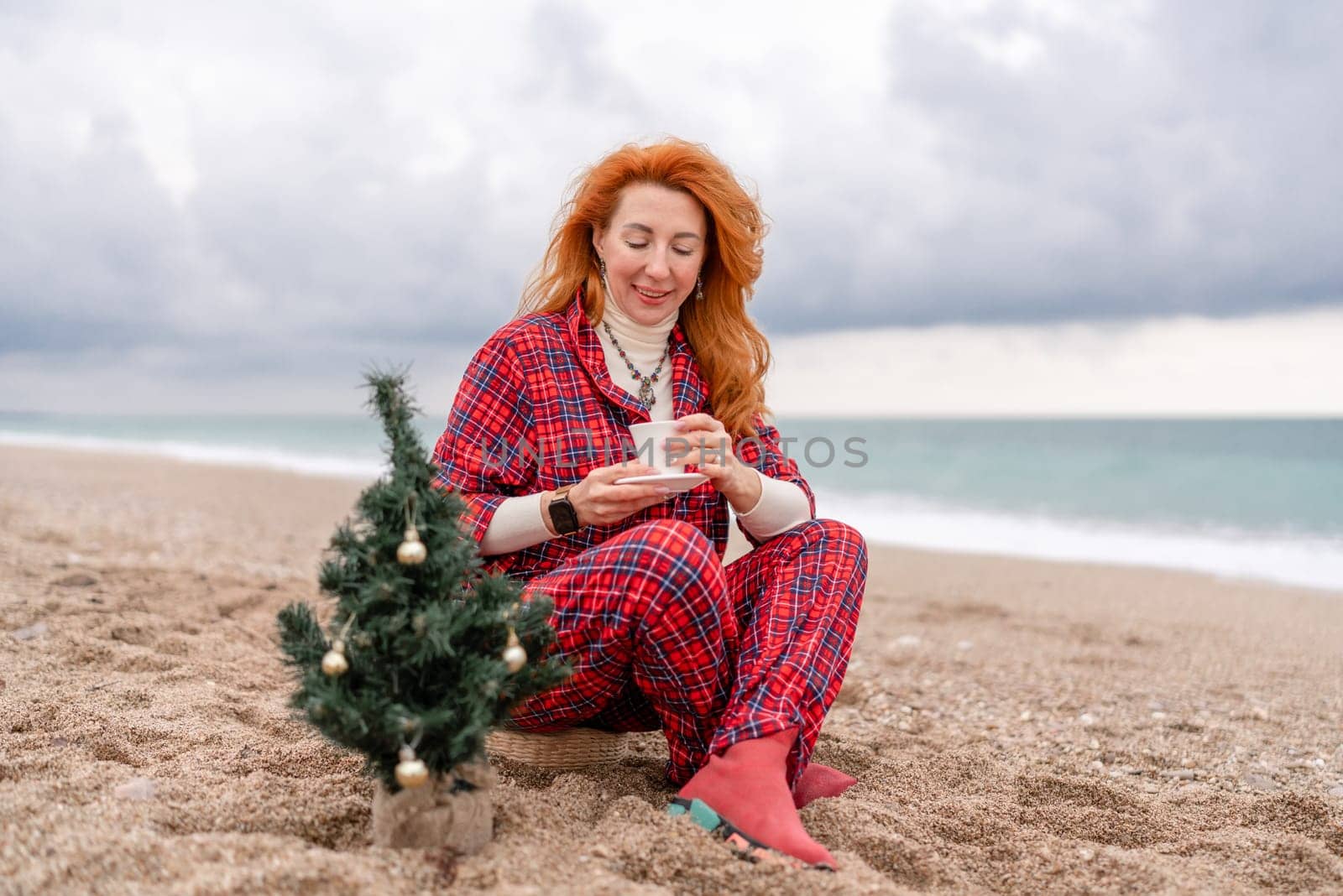 Sea Lady in plaid shirt with a mug in her hands enjoys beach with Christmas tree. Coastal area. Christmas, New Year holidays concep.