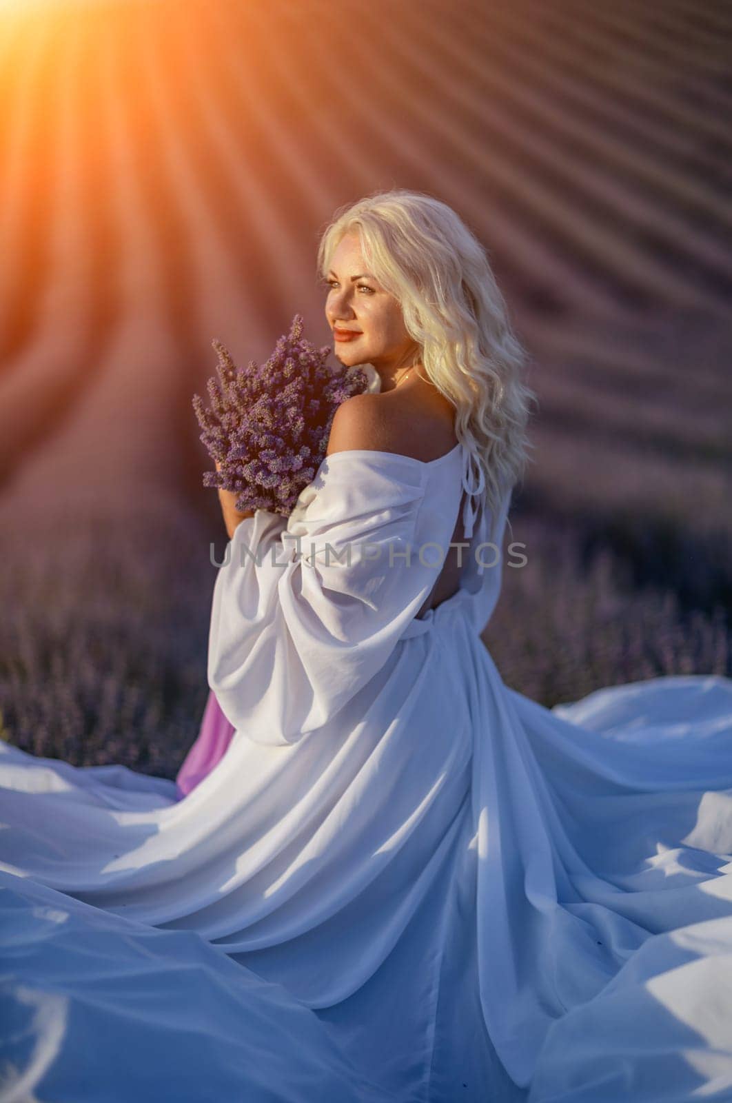 Blonde woman poses in lavender field at sunset. Happy woman in white dress holds lavender bouquet. Aromatherapy concept, lavender oil, photo session in lavender by Matiunina