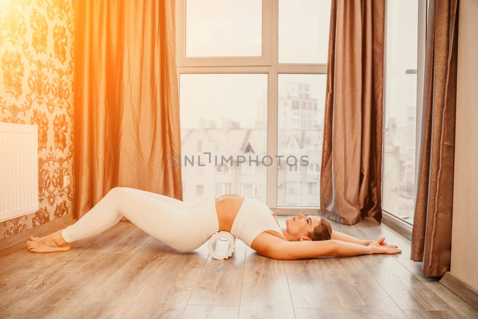 Adult athletic woman, in white bodysuit, performing fascia exercises on the floor - caucasian woman using a massage foam roller - a tool to relieve tension in the back and relieve muscle pain - the concept of physiotherapy and stretching training.