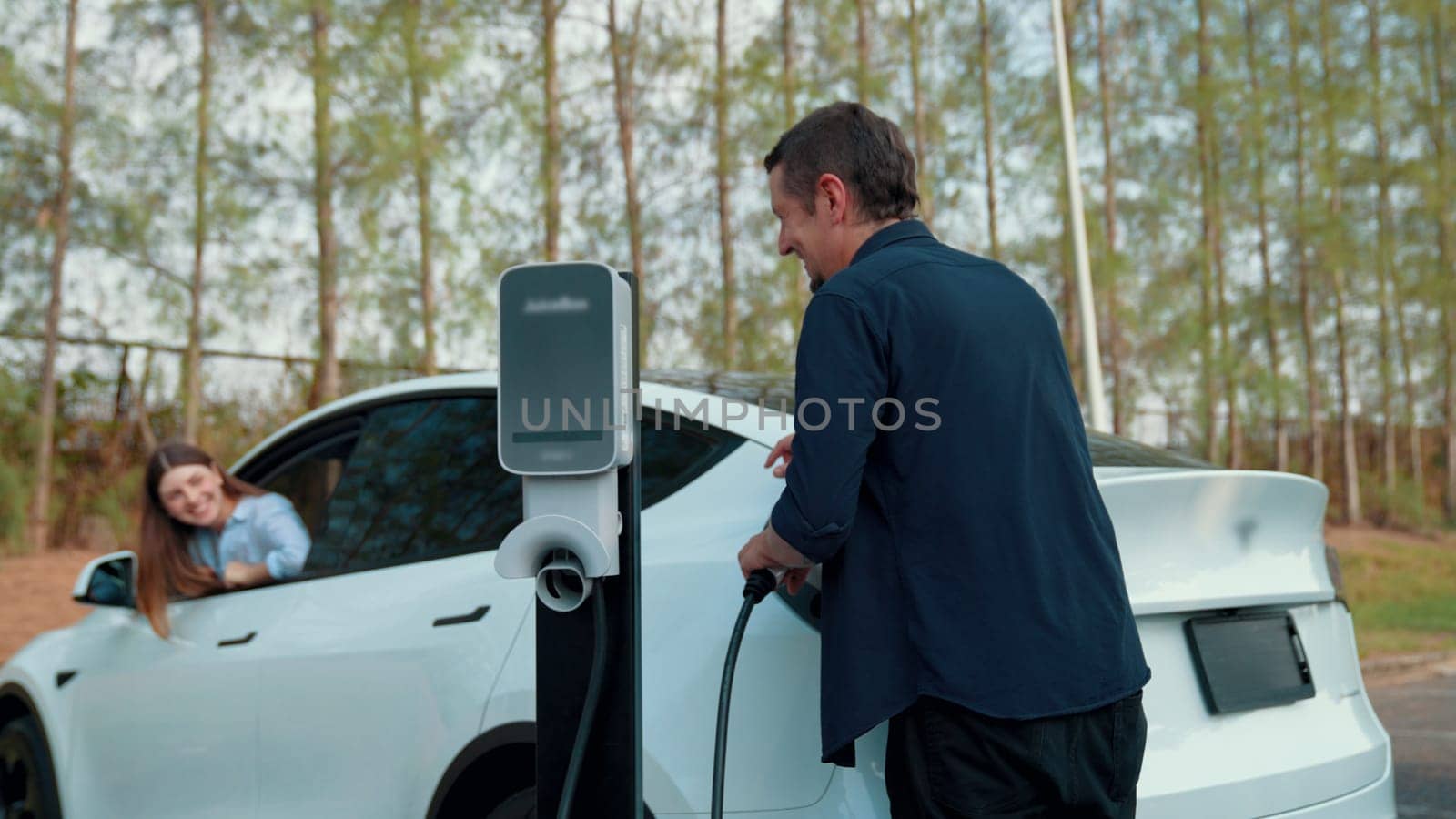 EV car recharging electricity for battery by lovey couple traveling during their road trip travel by eco friendly electric vehicle in national park autumnal forest on holiday. Exalt