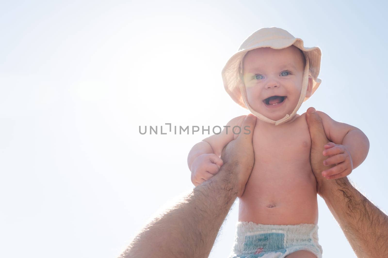 Baby held high against a sunny backdrop by a loving father. Concept of joy and freedom in early childhood by Mariakray