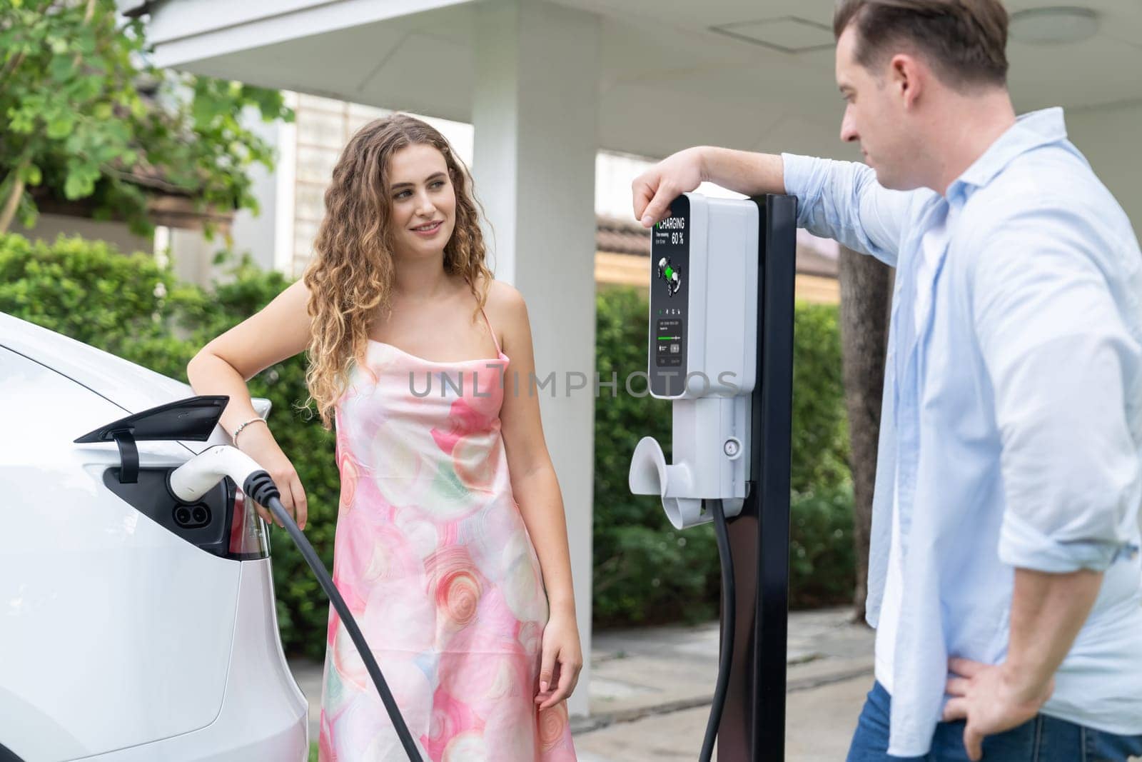 Happy and lovely couple with eco-friendly conscious recharging electric vehicle from EV home charging station. EV car technology utilized for residential home to future sustainability. Synchronos