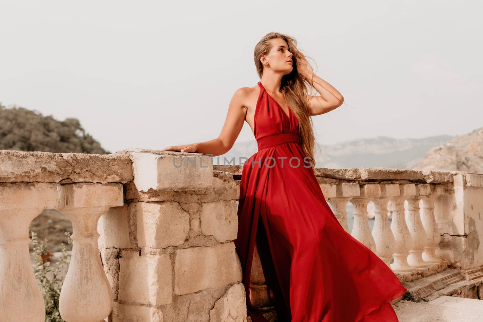 Side view a Young beautiful sensual woman in a red long dress posing on a volcanic rock high above the sea during sunset. Girl on the nature on overcast sky background. Fashion photo