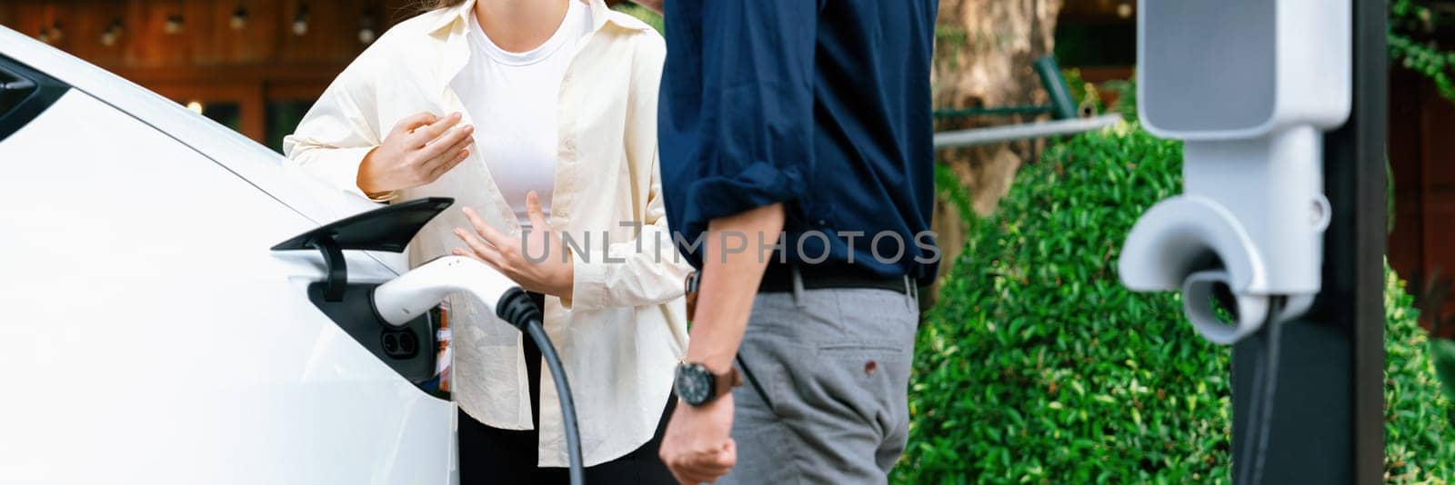 Young couple recharge electric car battery from charging station in green city park in springtime. Rechargeable EV car for sustainable environmental friendly urban travel lifestyle. Panorama Expedient