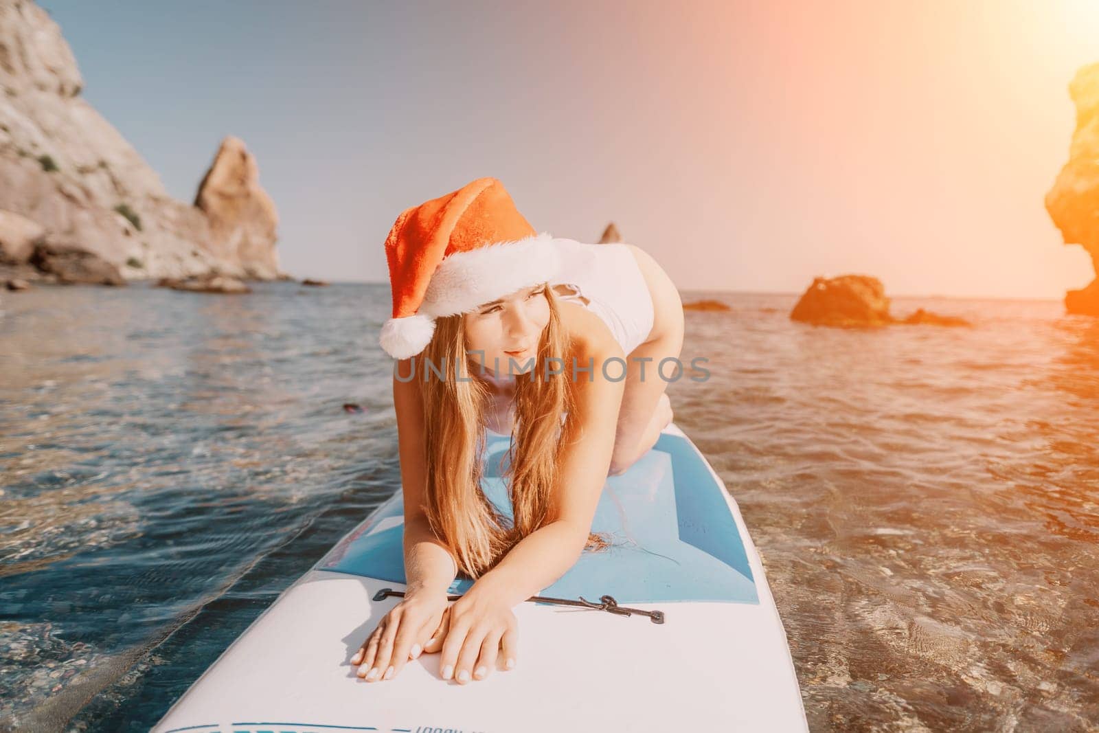 Woman sea sup. Close up portrait of happy young caucasian woman with long hair in Santa hat looking at camera and smiling. Cute woman portrait in a white bikini posing on sup board in the sea by panophotograph