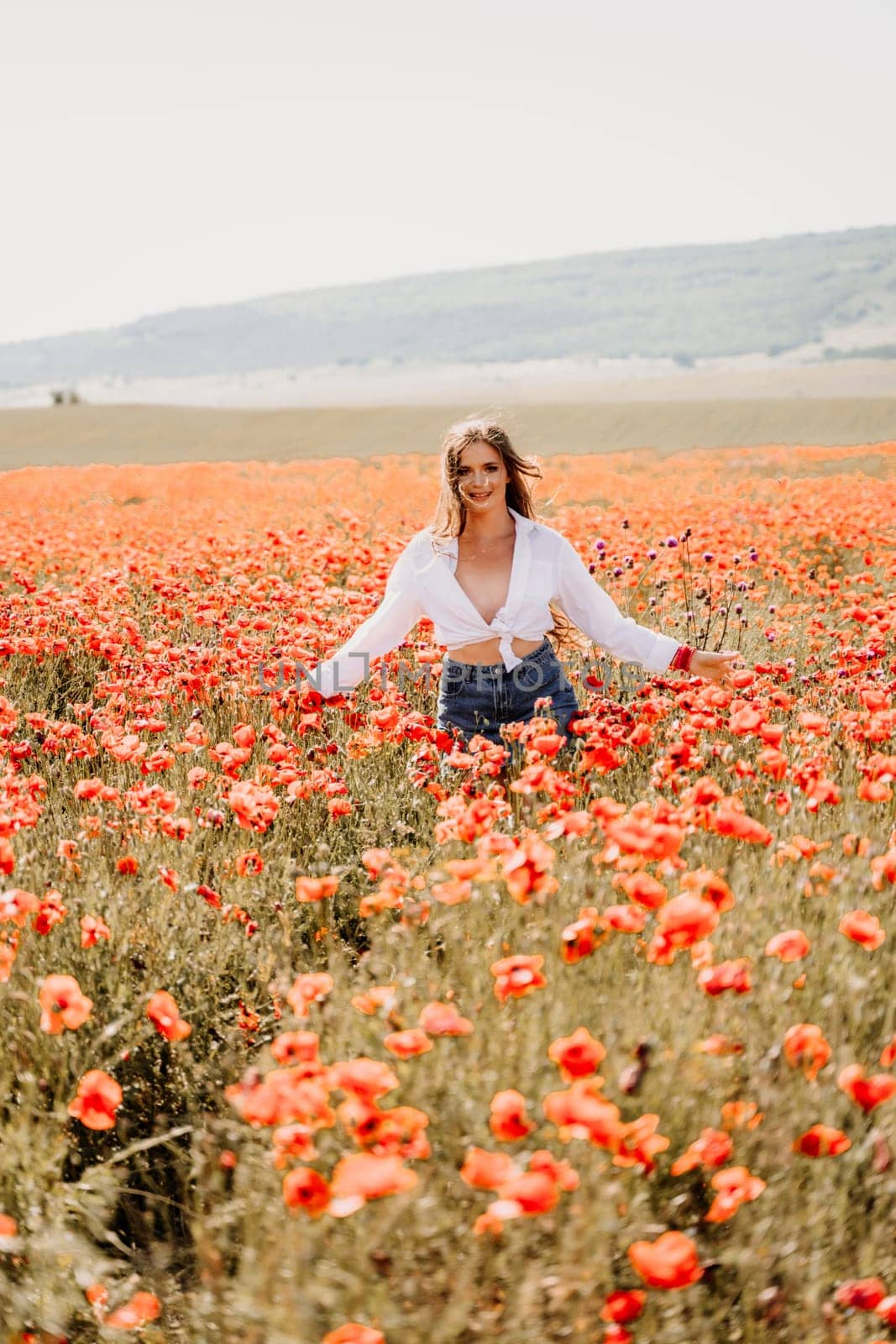 Happy woman in a poppy field in a white shirt and denim skirt with a wreath of poppies on her head posing and enjoying the poppy field