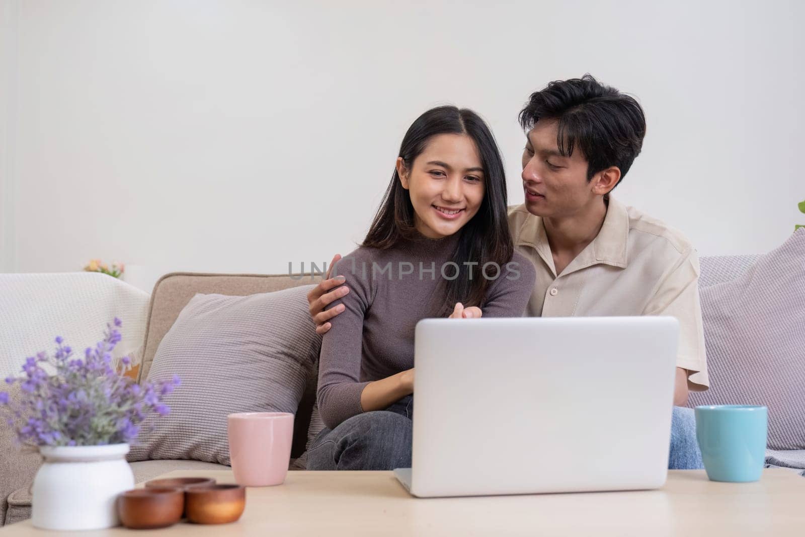 Asian couple watching movies on the internet using laptop at home Smiling Thai man and woman sitting on sofa, hugging each other, looking at computer screen. Surf the web together copy space.