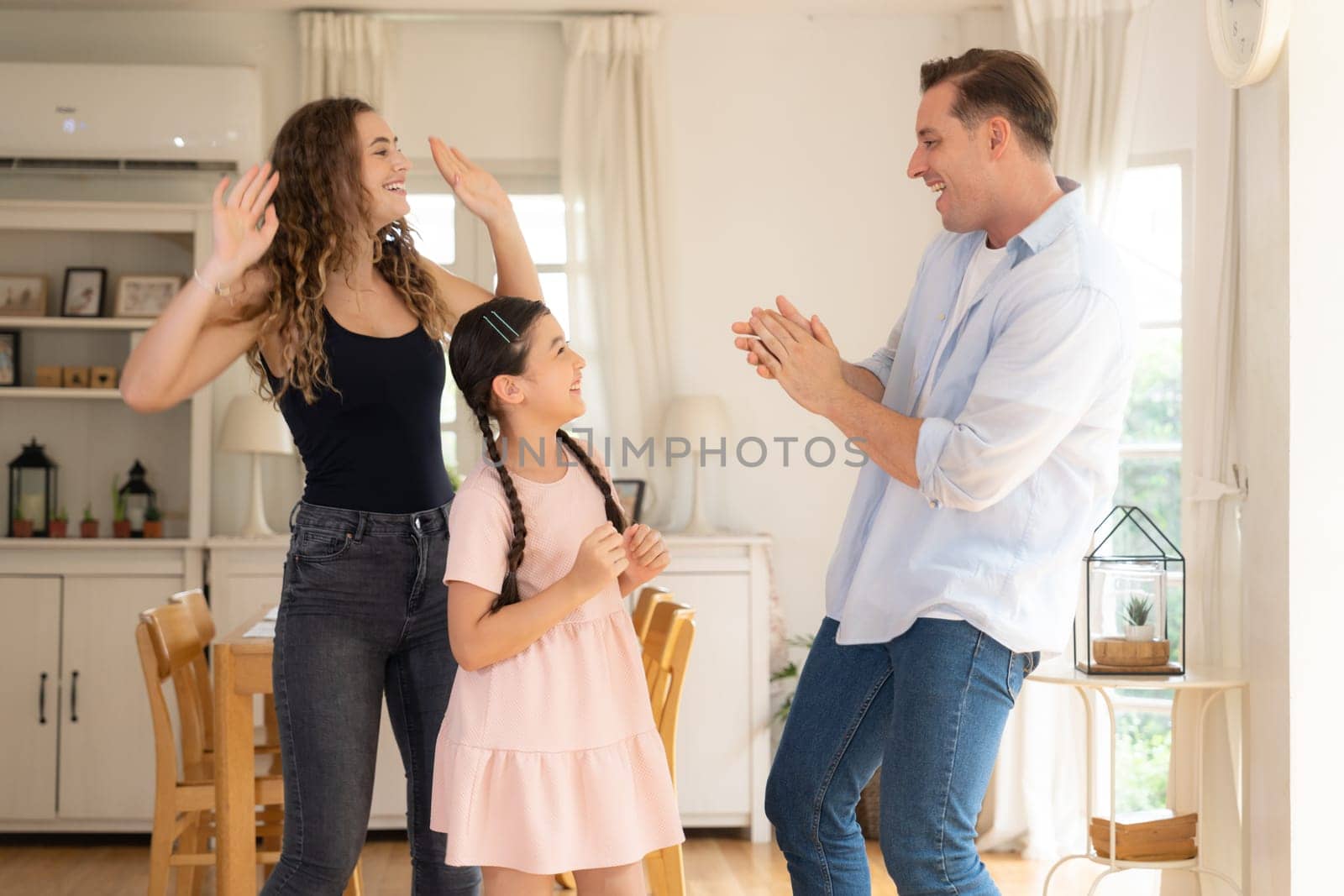 Happy family portrait with lovely little girl smiling and looking at camera, lovely and cheerful parent and their daughter sitting together in living room at home with warm daylight. Synchronos