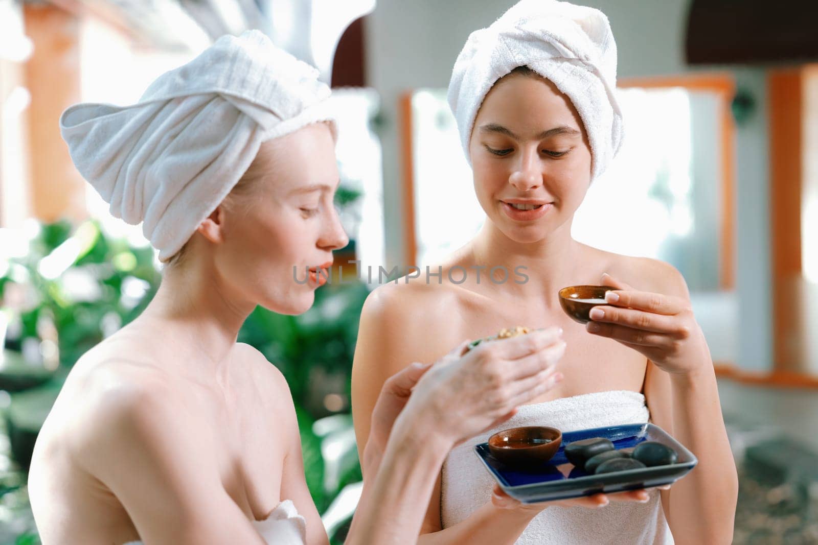 Couple of beautiful woman interested in homemade facial masks while sitting at spa salon. Attractive woman in white towel enjoy herbal masks with her friend surrounded by nature. Tranquility.