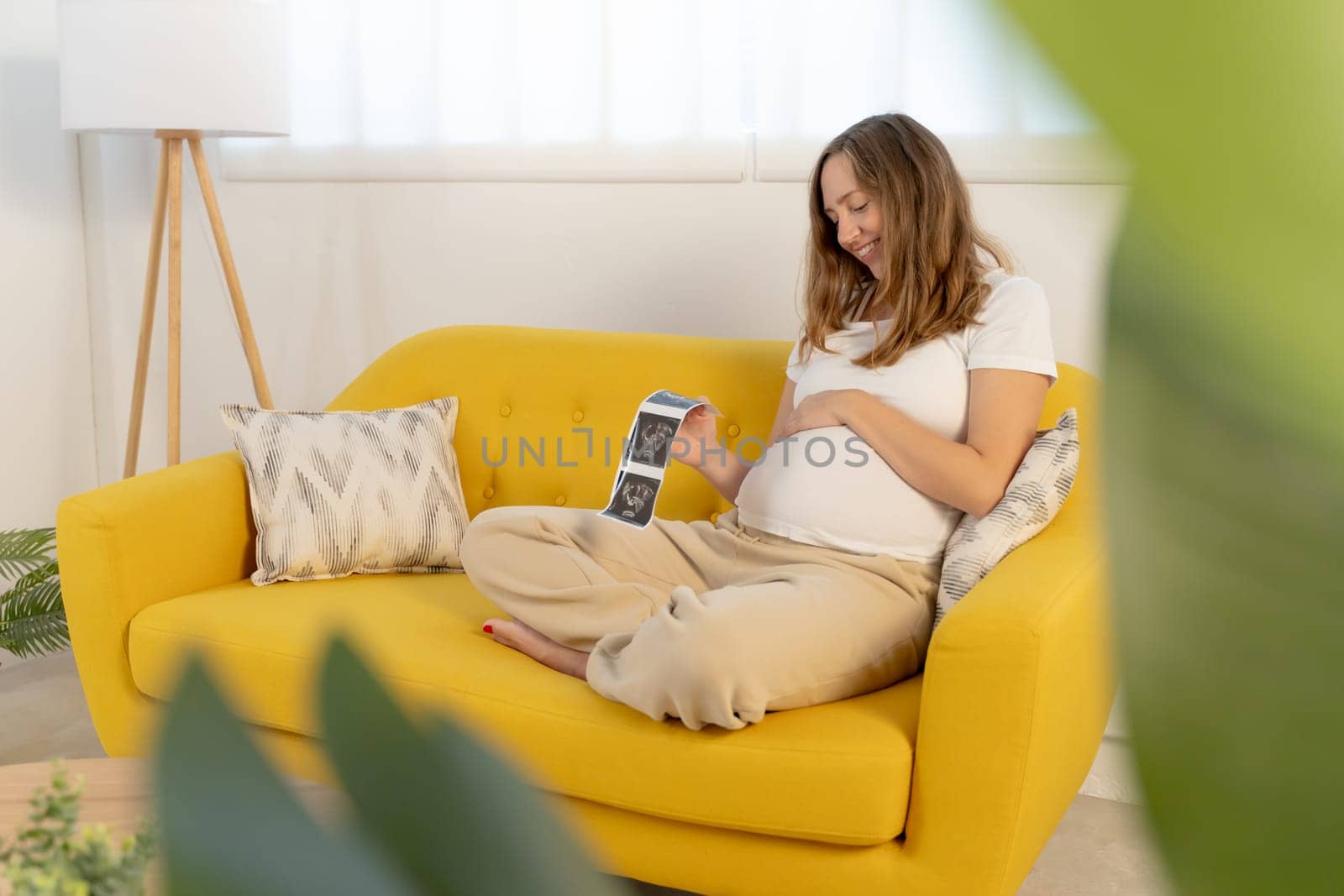 Pregnancy mother looking picture of ultrasound film and touching her abdomen. Woman checking baby result sitting relaxed and rest on sofa in living room at home. High quality photo