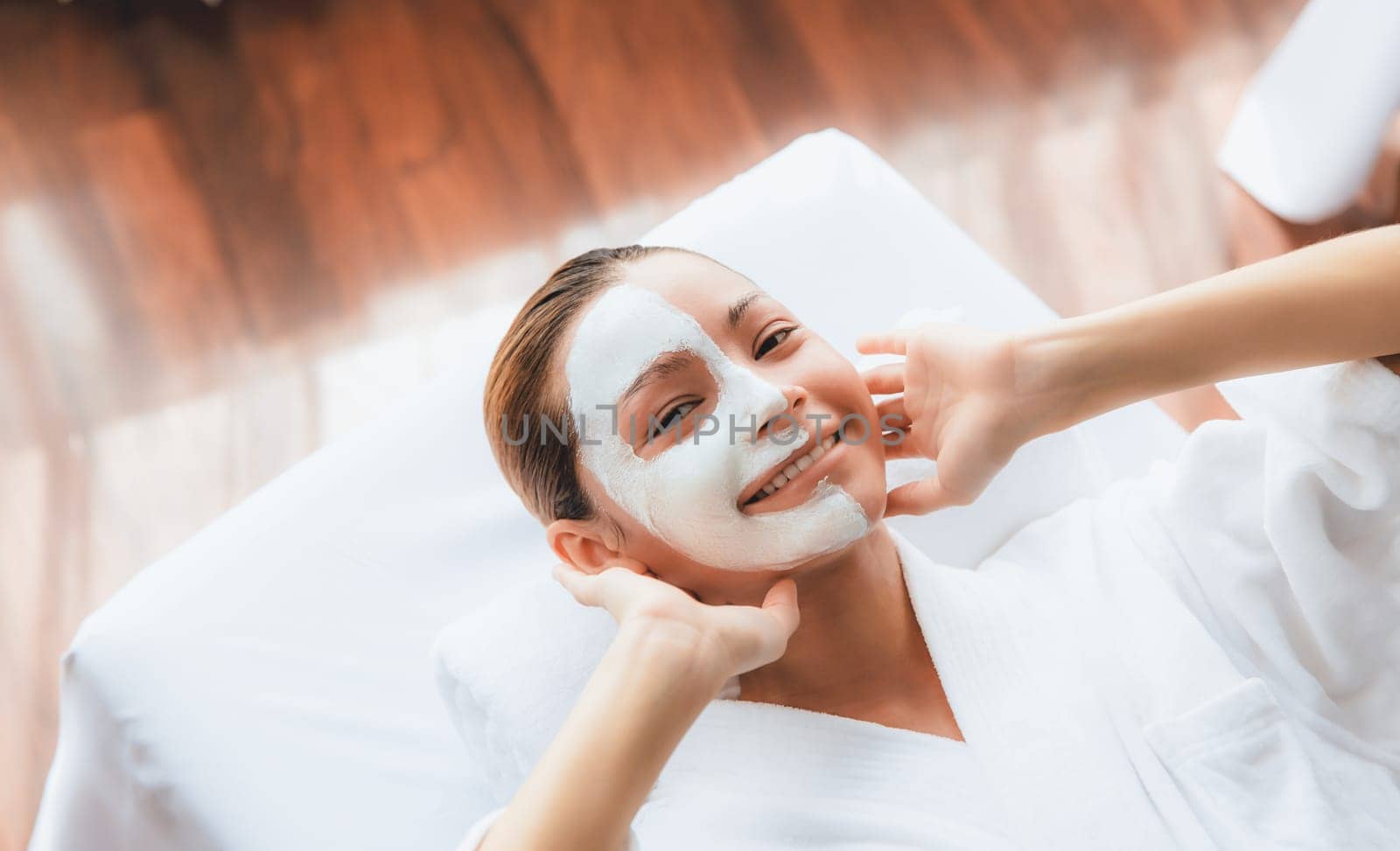 Serene daylight ambiance of spa salon, top view woman lying on massage table smiling and rejuvenating with face cream spa massage. Facial skin spa treatment and beauty care concept. Quiescent