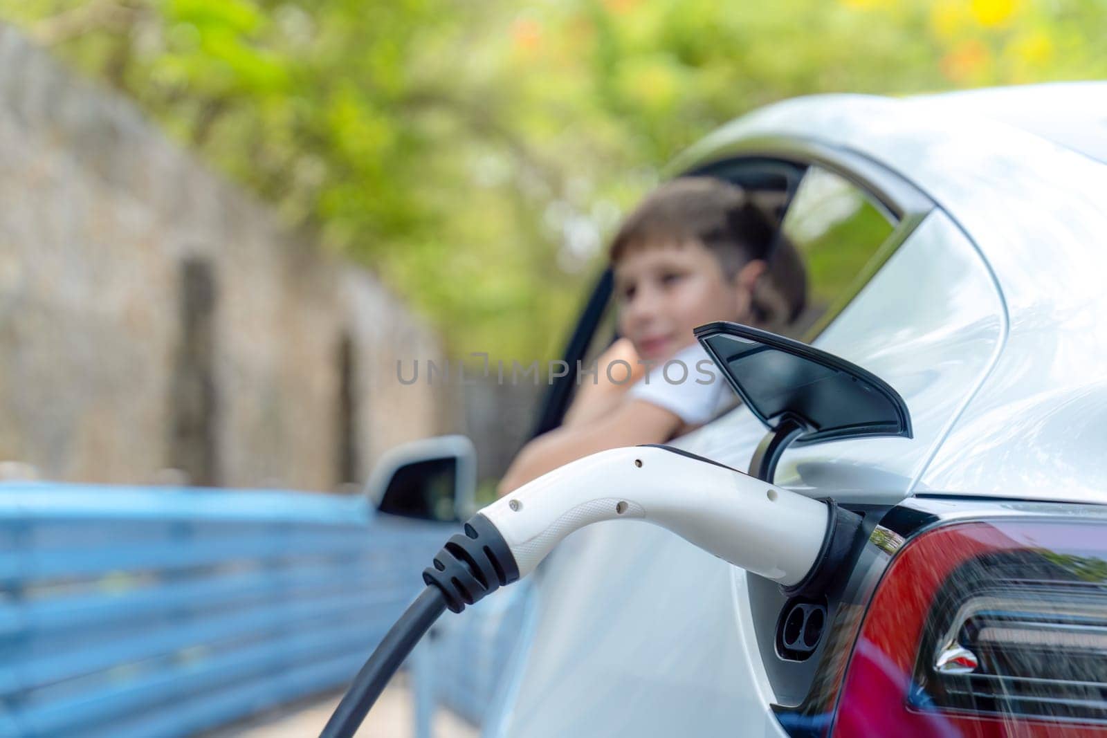 EV car recharge battery from EV charging station with blur little boy waiting inside the car in background. Road trip travel by alternative vehicle powered by clean and sustainable energy. Perpetual