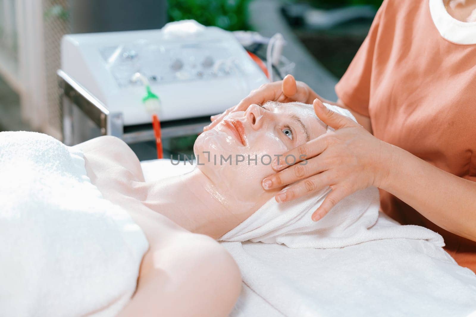 Portrait of beautiful caucasian woman having facial massage with homemade facial mask while lies on spa bed surrounded by beauty electrical equipment and peaceful nature environment. Tranquility.