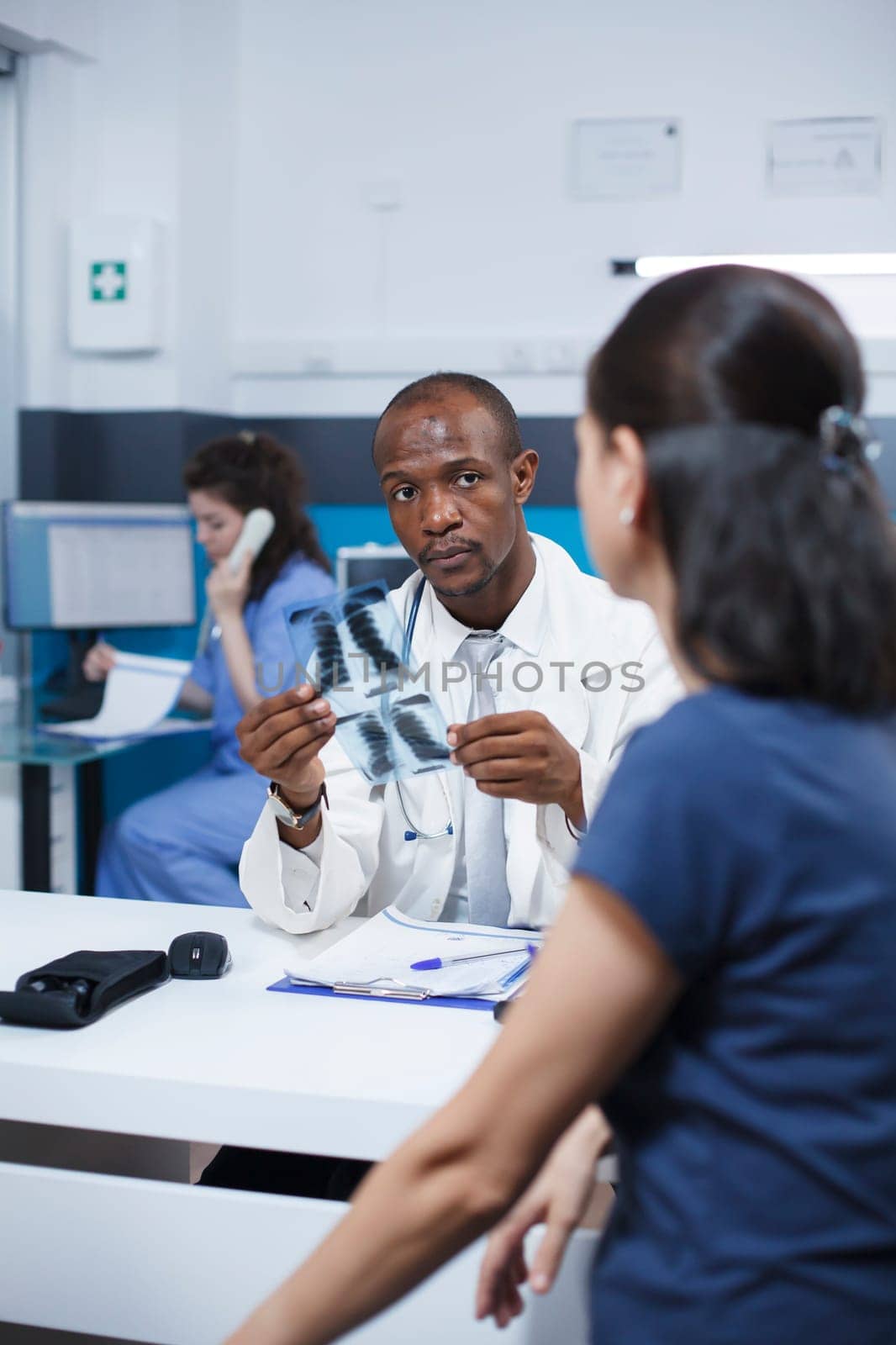 African American female specialist reviewing x-ray image and analyzing imaging results. Radiology expert examines radiography scan and provides diagnostic results to caucasian woman patient.