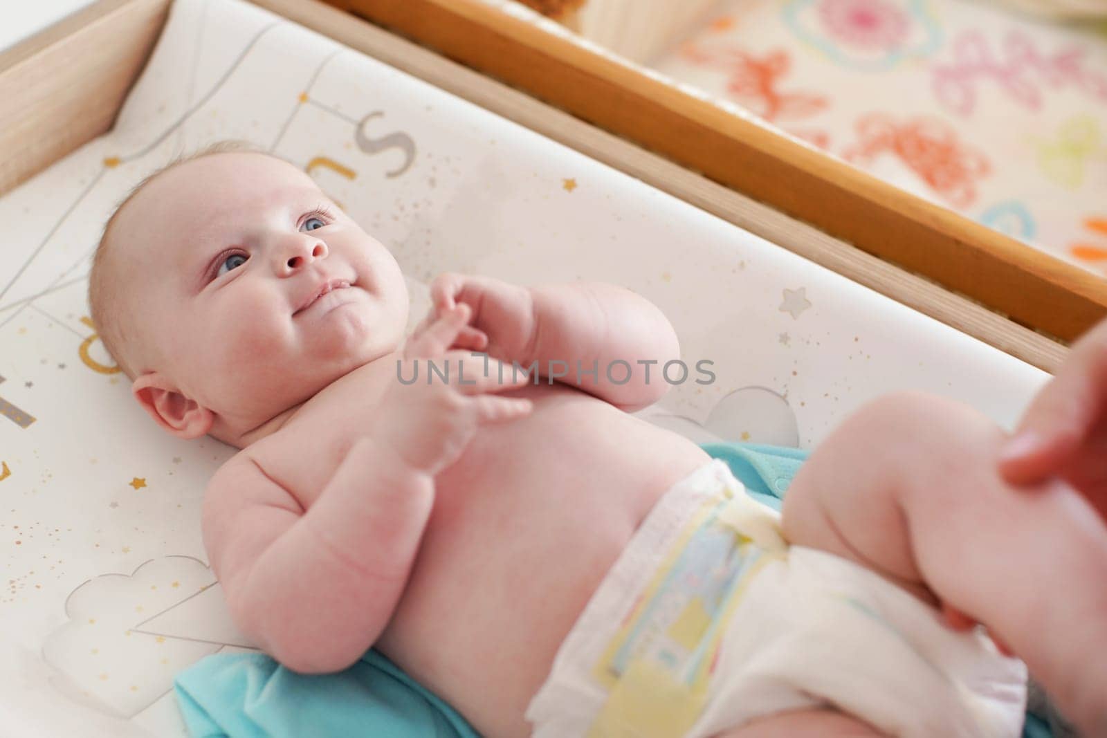 4 months old infant boy laying on changing desk, mother hands holding his legs by Ivanko