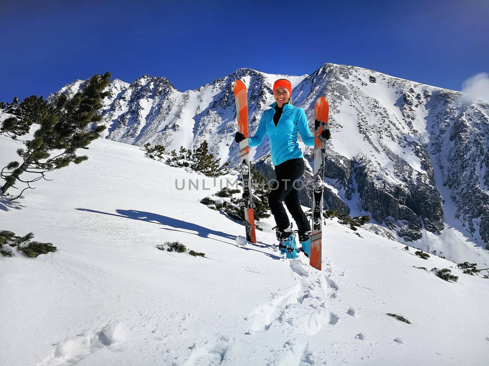 Young woman in light blue jacket standing, smiling happy holding with mountaineering ski and poles in hands, sun shines on snow, bright sky over mountains behind