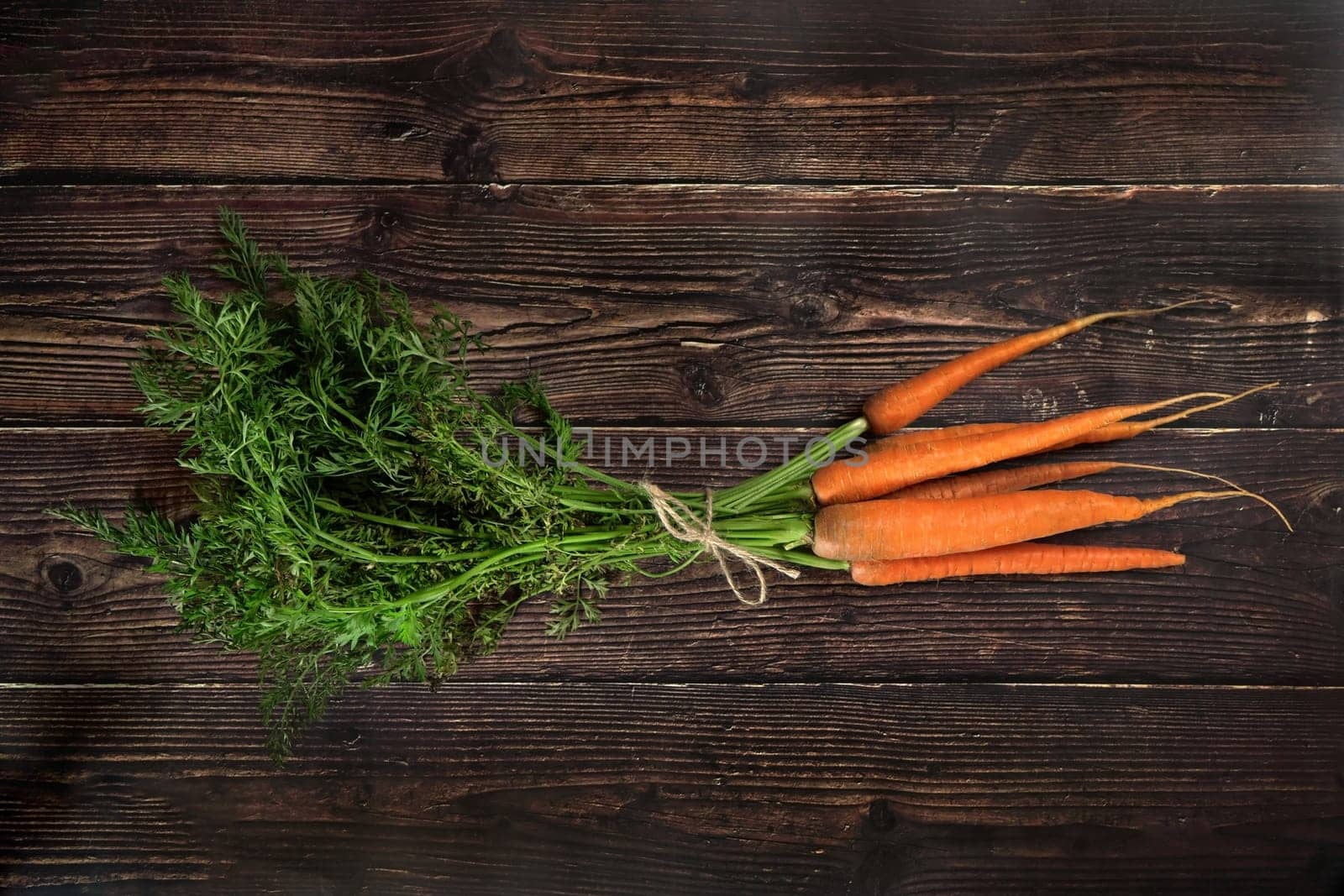 Top down view, bunch of fresh carrot roots with green leaves, holding together with rope cord, on dark wooden board