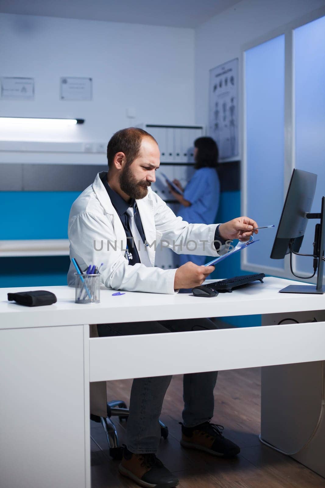Detailed image of male doctor at the clinic desk going over and analyzing his notes. Caucasian man utilizing desktop computer and clipboard as he prepares for patient medical consultations.