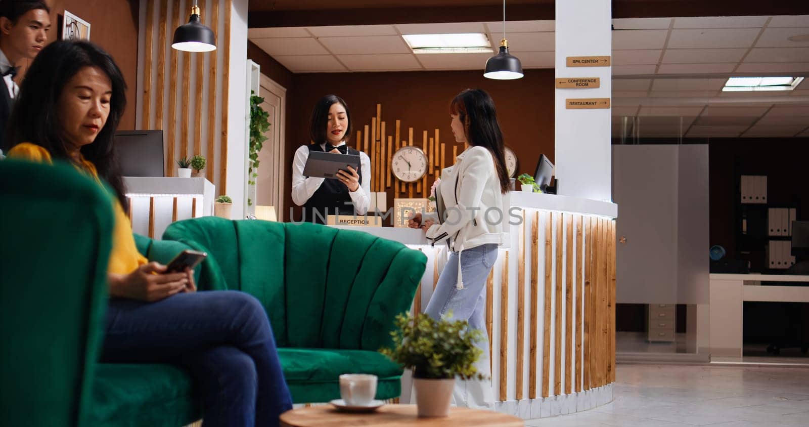 Asian guest signing check in papers on tablet at reception, registering her room confirmation while bellboy is helping with her luggage. Five star hotel staff team friendly welcoming client.