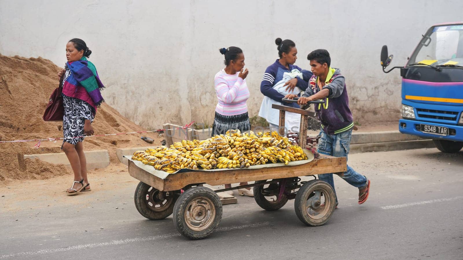 Antananarivo, Madagascar - April 24, 2019: Unknown Malagasy man, pulling cart with ripe bananas on main asphalt road, other women, and car in background. Bananas are cheap and popular at Madagascar by Ivanko