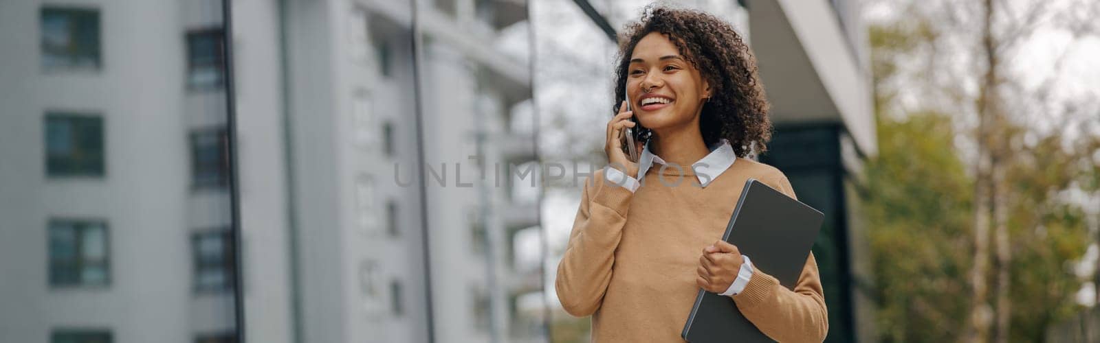 Pretty female entrepreneur is talking phone while standing with laptop on modern building background by Yaroslav_astakhov