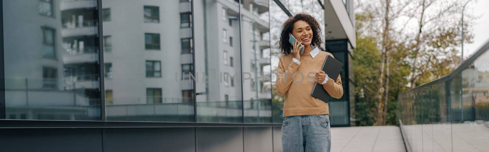 Pretty female entrepreneur is talking phone while standing with laptop on modern building background