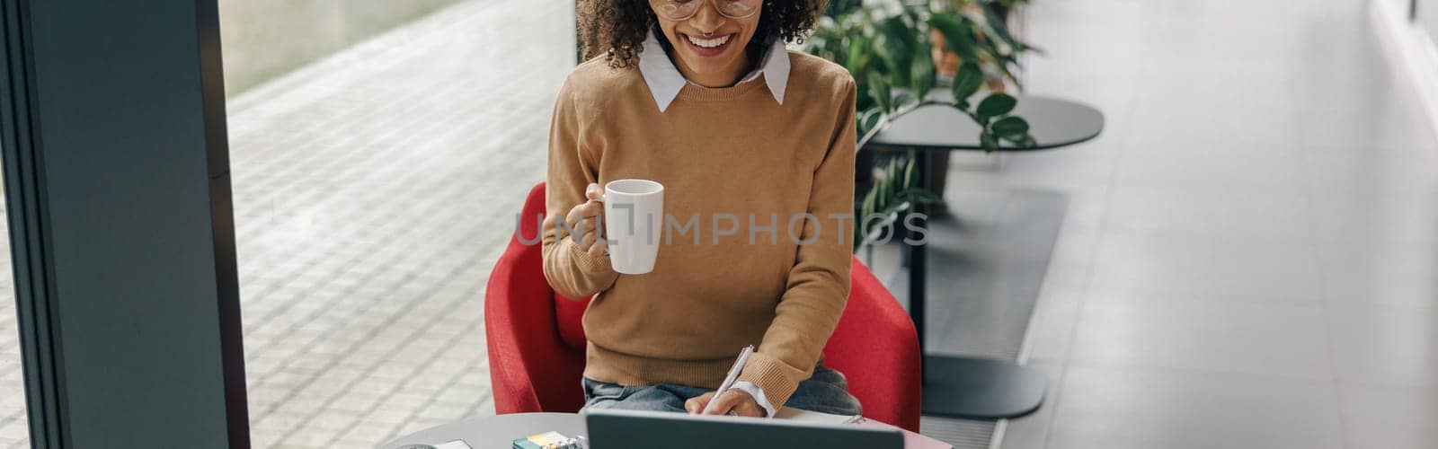 Smiling woman entrepreneur working on laptop, making notes in cozy coworking space interior by Yaroslav_astakhov