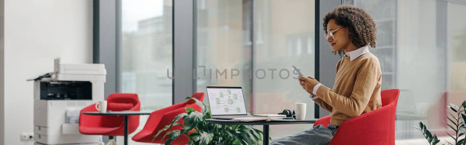 Stylish business woman is use phone while working on laptop in modern office near window