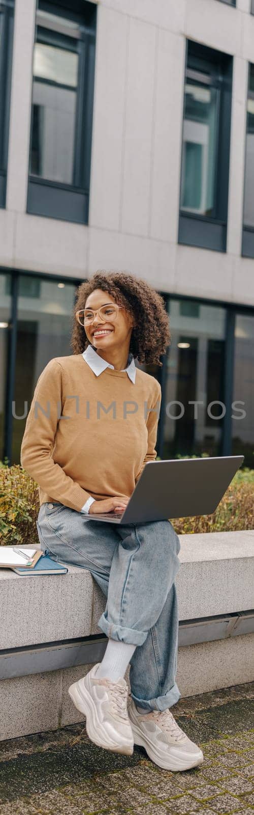Smiling female freelancer working laptop while sitting outside on background of office building