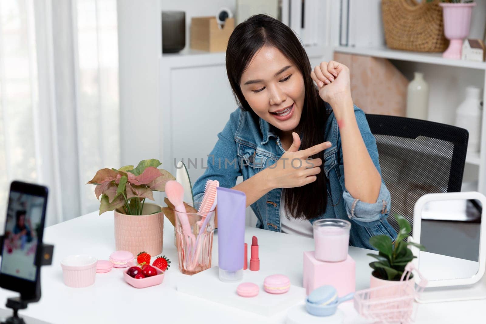 Young beautiful Asian showing cosmetic makeup lipstick matte pastel color testing in bestseller collection on arm test promotion on social media online recording smartphone at modern room. Stratagem.