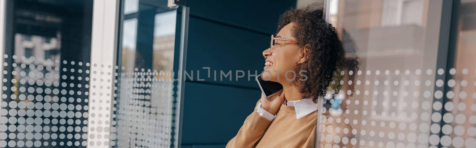 Smiling businesswoman is talking phone with client while standing on modern office background