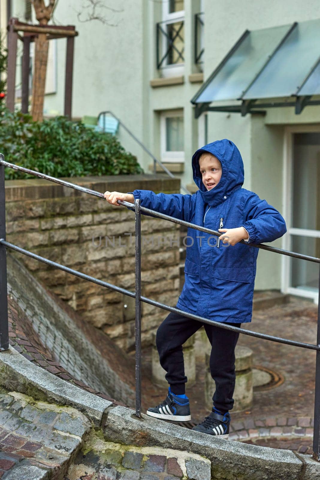 Joyful Boy in Blue Winter Jacket on Outdoor Stairs with City Buildings in the Background, Autumn or Winter. Experience the delight of the season with this heartwarming image featuring a cheerful 8-year-old boy wearing a blue winter jacket and hood, standing on an outdoor staircase, holding onto the railing. The photograph captures the vibrant spirit of autumn or the serene beauty of winter against the backdrop of city buildings.