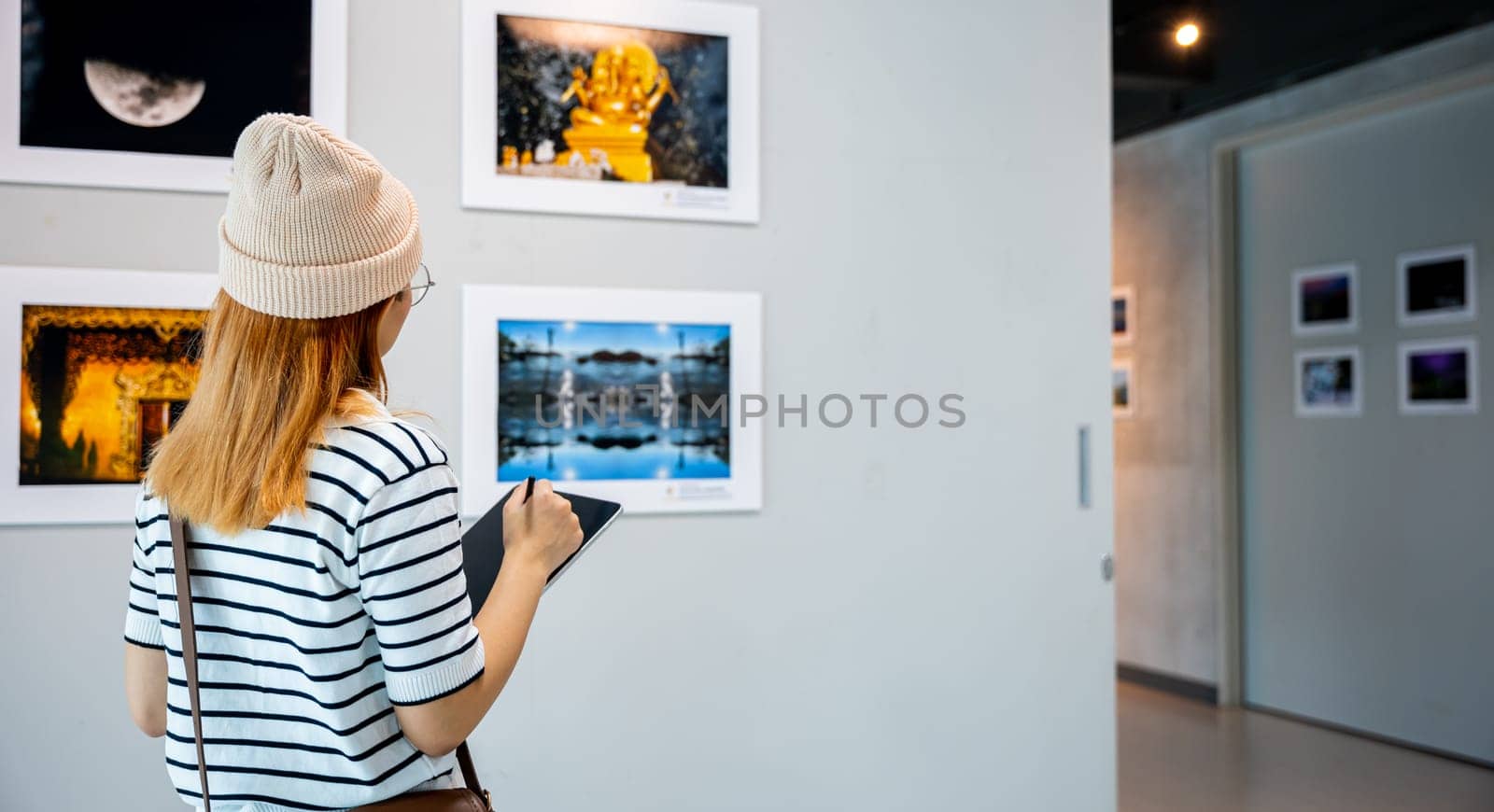 Young person at photo frame write digital book leaning against at show exhibit artwork gallery, Asian woman holding tablet at art gallery collection in front framed paintings looking pictures on wall