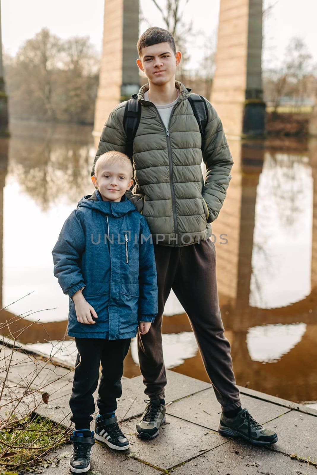 Winter Riverside Moment: Two Boys (8 and 17 Years Old) in Jackets Stand by the River and Bridge Piers, Autumn or Winter. Capture the essence of seasonal camaraderie with this image featuring two boys, aged 8 and 17, donned in winter jackets, standing against the backdrop of a river and bridge piers. The photograph beautifully encapsulates the spirit of autumn or winter, showcasing the bond between the boys in a serene riverside setting.